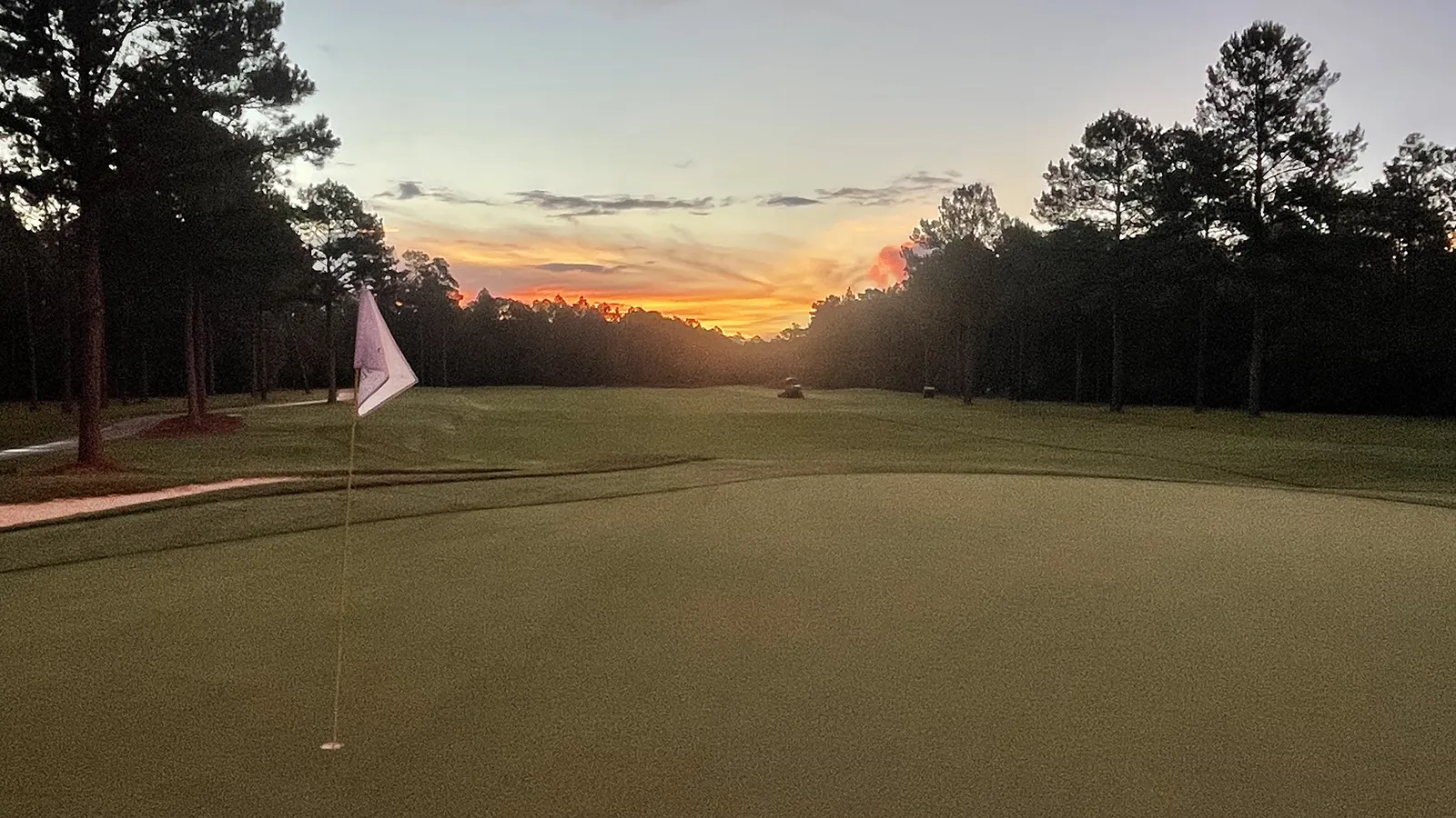 A green at Grand Bear Golf Club at dusk, with a flagstick in the foreground and an orange sunset behind the pine treeline